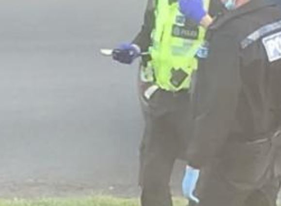 A police officer wearing gloves is seen holding an object, which could be a machete, outside the garden where the machete was discovered