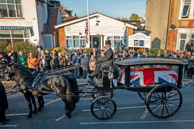 Members of the public pay their respects as the horse drawn hearse carrying the coffin of Sir David Amess, arrives at his constituency office at Iveagh Hall, in Leigh-on-Sea, following his funeral service. Picture date: Monday November 22, 2021..